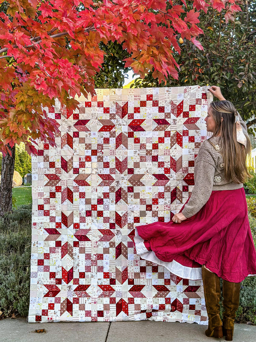 Maple Red Patchwork Low Volume Fabric Star Quilt Made from Maple Sonnet Quilt Kit with woman in knit sweater and red dress in front of red autumn leaves. 