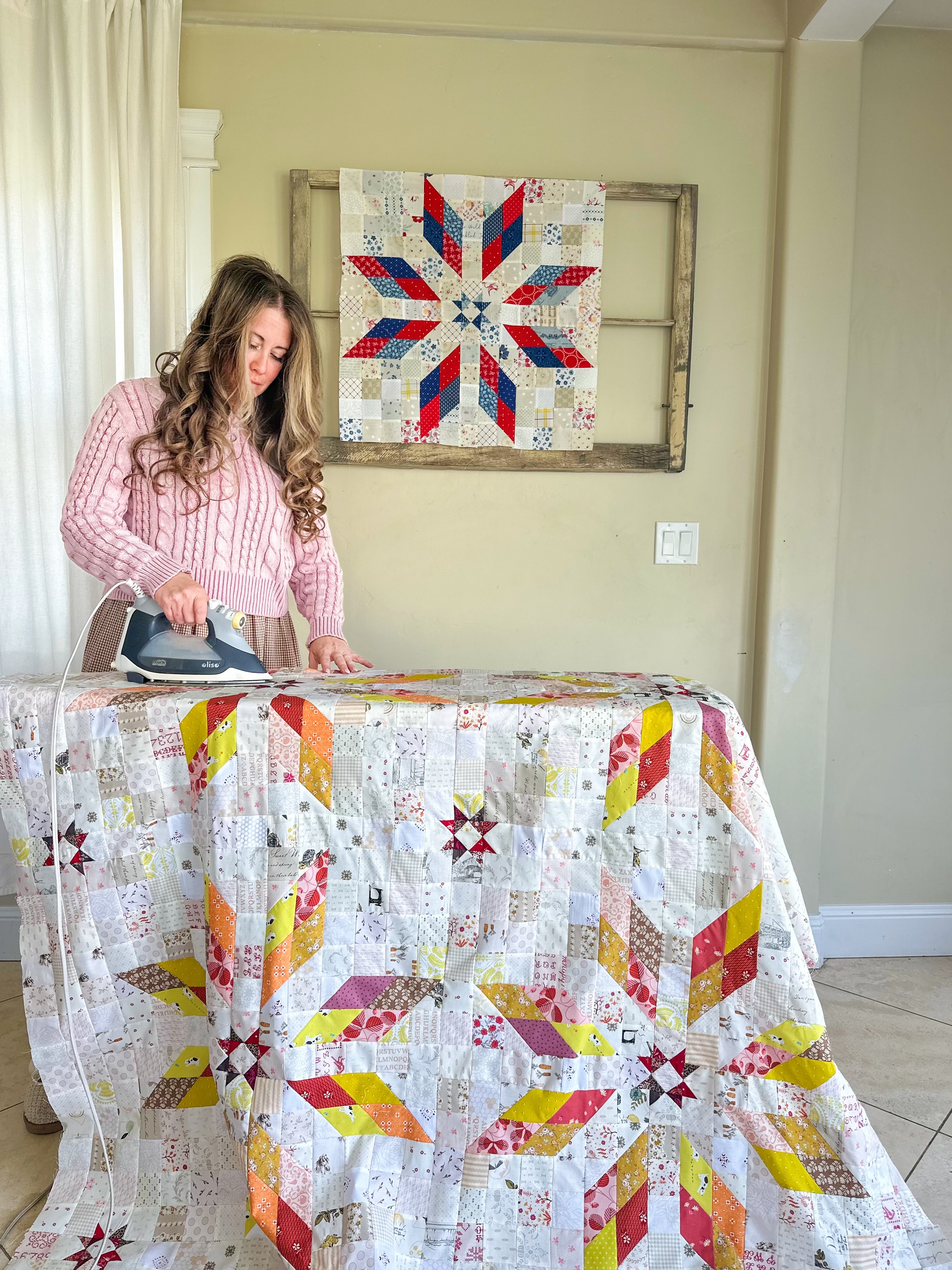 woman ironing a quilt with a red white and blue quilt block on the wall behind her. 